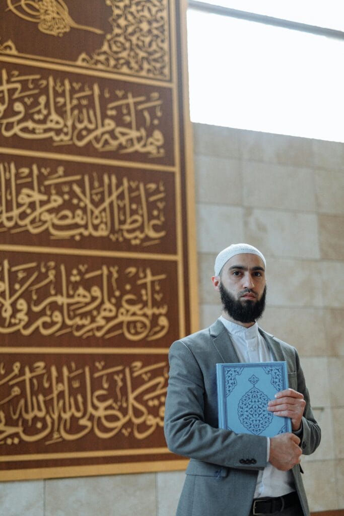pexels-photo-8489316-8489316 A bearded Muslim man in a suit holds a Quran inside a mosque with Arabic calligraphy on the wall.