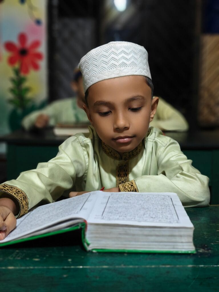 pexels-photo-37350650-37350650 A young boy in traditional attire reads the Quran attentively at a classroom desk.